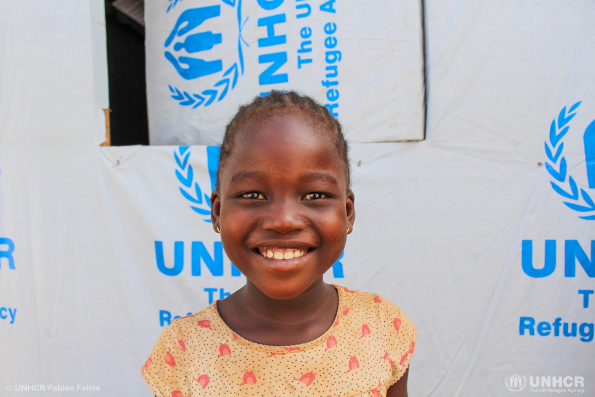 sisé smiles at the camera in front of a UNHCR shelter