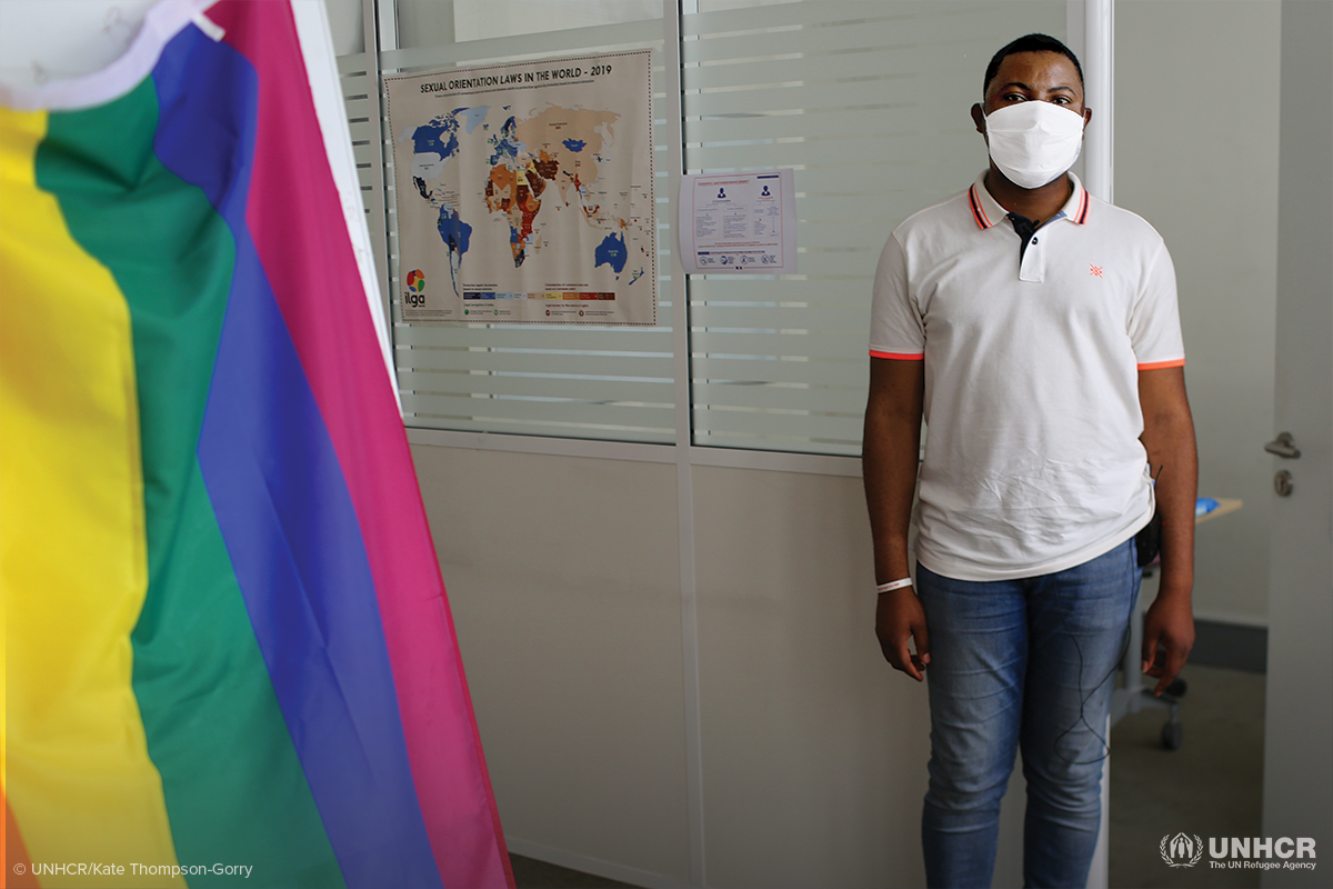 thomas stands in an office with a mask on next to a rainbow flag